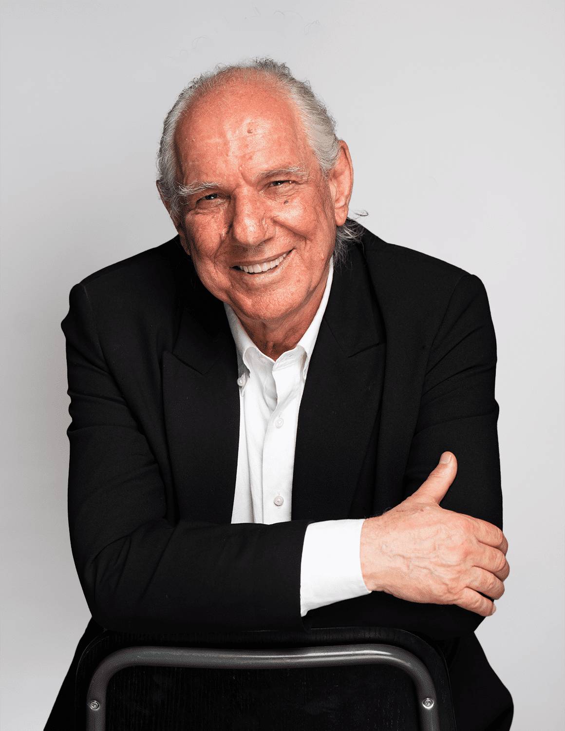Smiling elderly man in a black suit with folded arms.